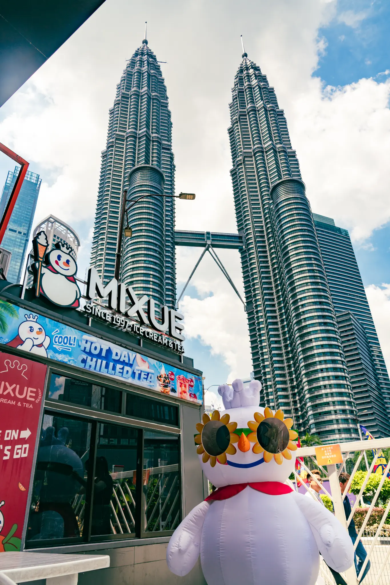 MIXUE store with snowman mascot wearing sunflower sunglasses in front of Petronas Towers Malaysia